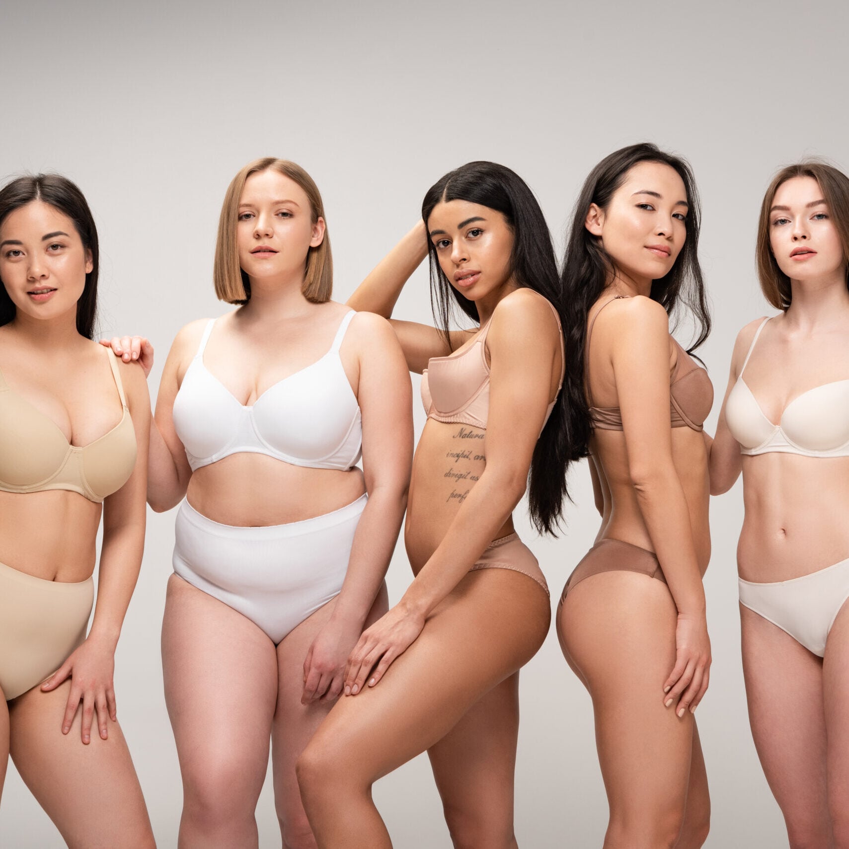 five smiling multiethnic girls smiling and posing at camera isolated on grey, body positivity concept